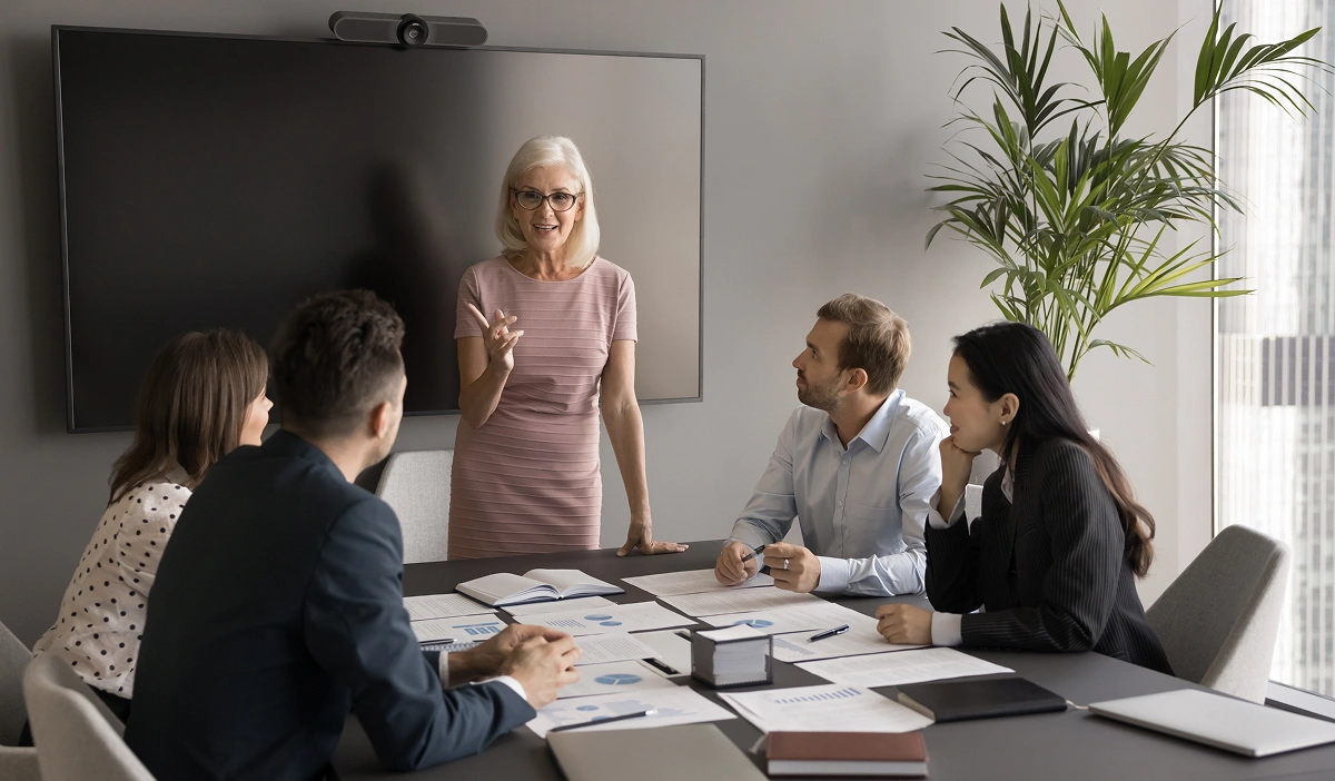 Senior woman in pink dress speaks at a conference table as four colleagues listen, papers spread across the table.
